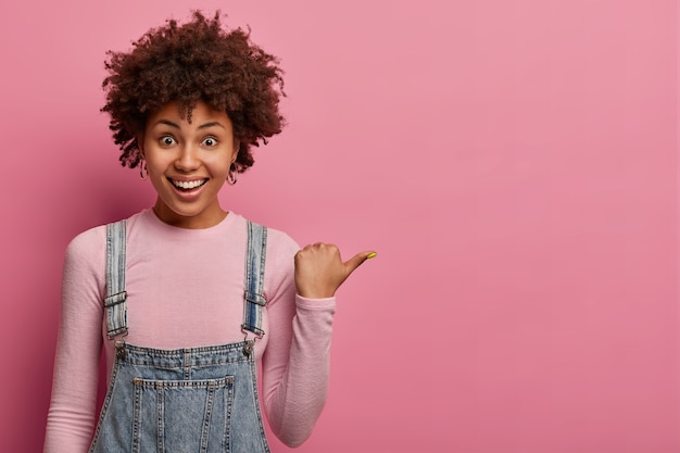 La niña afroamericana positiva señala con el pulgar, demuestra el espacio de la copia en la pared rosa en blanco, tiene una expresión feliz y amistosa, se viste informalmente, posa en el interior, sugiere ir a la derecha, dice seguir esta dirección