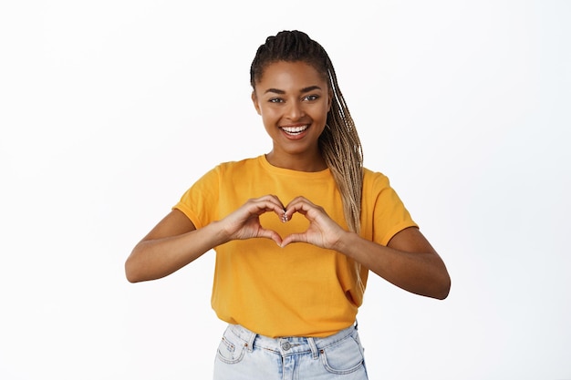 Niña afroamericana sonriente muestra un gesto de amor al corazón y se ve linda a la cámara confesión romántica de pie sobre fondo blanco