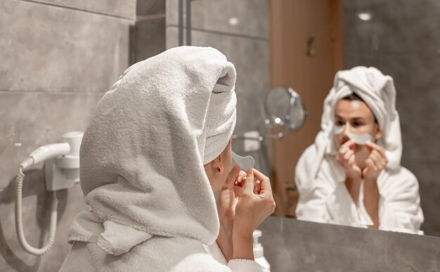 Una niña un albornoz y con una toalla en la cabeza se pega parches debajo de los ojos en el baño frente al espejo.