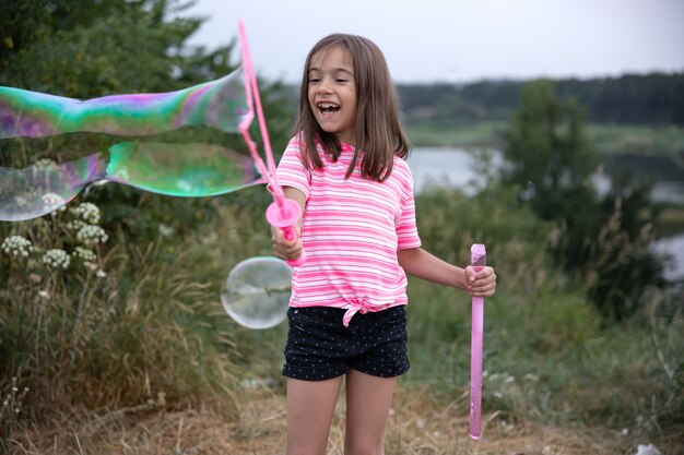 Niña alegre juega con grandes pompas de jabón en la naturaleza.