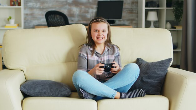 Niña alegre jugando videojuegos con controlador inalámbrico sentado en el sofá.