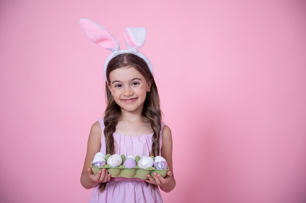 Niña alegre con orejas de conejo de Pascua y una bandeja de huevos en sus manos en un estudio rosa