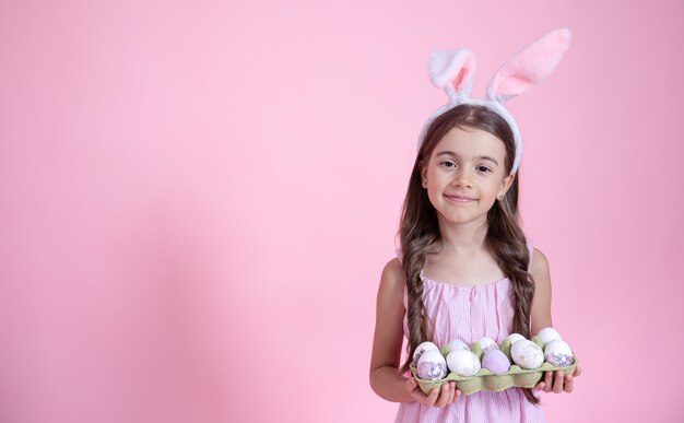 Niña alegre con orejas de conejo de Pascua y una bandeja de huevos en sus manos sobre una pared rosa. Concepto de vacaciones de semana Santa.