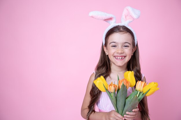 Niña alegre con orejas de conejo de Pascua sonríe y sostiene un ramo de tulipanes en sus manos en un estudio rosa