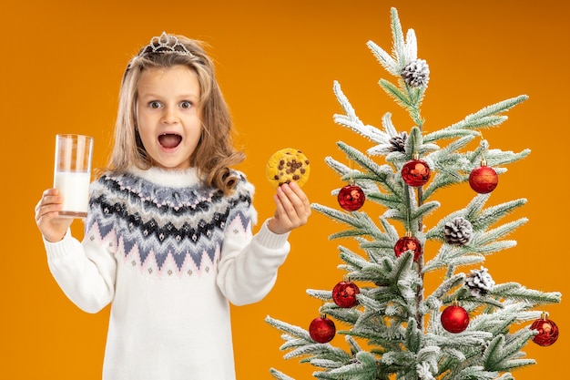 Foto gratuita niña alegre de pie cerca del árbol de navidad con tiara con guirnalda en el cuello sosteniendo un vaso de leche con galletas aislado sobre fondo naranja