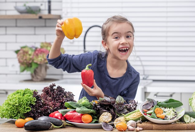Niña alegre sosteniendo pimiento sobre un fondo de diversas verduras. Concepto de comida sana.