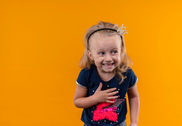 Foto gratuita una niña alegre vistiendo camisa azul marino en diadema de corona sonriendo mientras mira de lado en una pared naranja