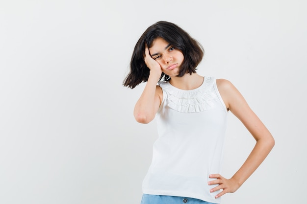 Niña apoyada en la palma de la mejilla en blusa blanca, pantalones cortos y mirando deprimido, vista frontal.