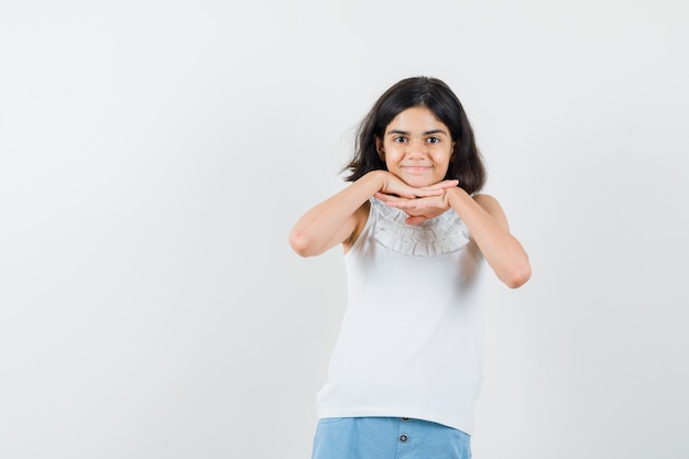 Niña apoyando la barbilla en las manos en blusa blanca, pantalones cortos y bonita. vista frontal.