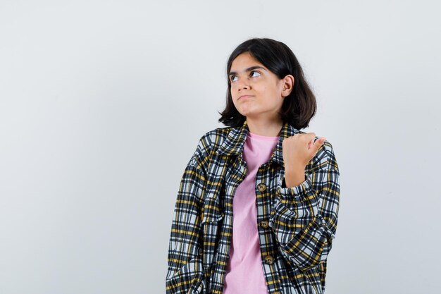 Niña apretando el puño y mirando a otro lado con camisa a cuadros y camiseta rosa y mirando pensativo, vista frontal.