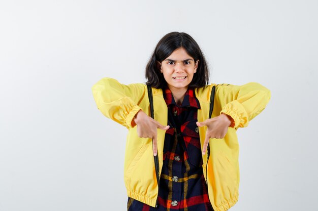 Niña apuntando hacia abajo en camisa a cuadros, chaqueta y mirando feliz