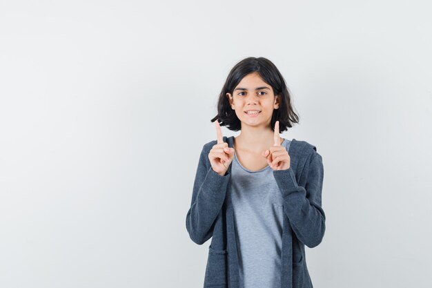 Niña apuntando hacia arriba en camiseta, chaqueta y luciendo alegre.