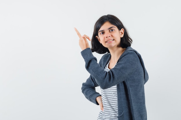 Niña apuntando hacia arriba en camiseta, chaqueta y luciendo orgullosa.