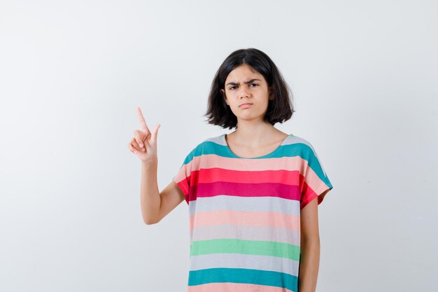 Niña apuntando hacia arriba, haciendo muecas en camiseta, jeans y luciendo disgustada. vista frontal.