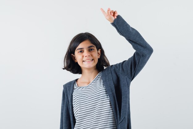 Niña apuntando hacia arriba levantando el brazo en camiseta, chaqueta y luciendo feliz.