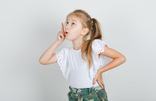 Niña apuntando hacia arriba con la mano en la cintura en camiseta blanca