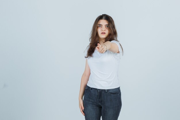 Niña apuntando a la cámara en camiseta, jeans y mirando sorprendido, vista frontal.