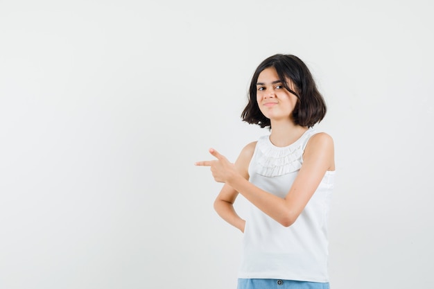 Niña apuntando hacia el lado en blusa blanca, pantalones cortos y mirando alegre, vista frontal.