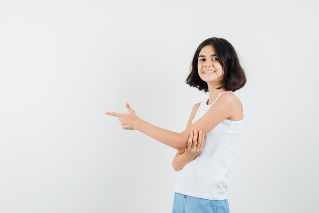 Niña apuntando hacia el lado en blusa blanca, pantalones cortos y mirando alegre, vista frontal.