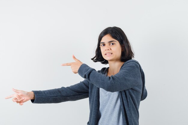 Niña apuntando a un lado en camiseta, chaqueta y mirando confiada.