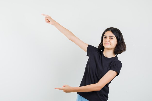 Niña apuntando a un lado en camiseta negra, pantalones cortos y mirando confiado, vista frontal.