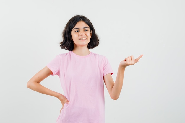 Niña apuntando a un lado en camiseta rosa y mirando optimista, vista frontal.