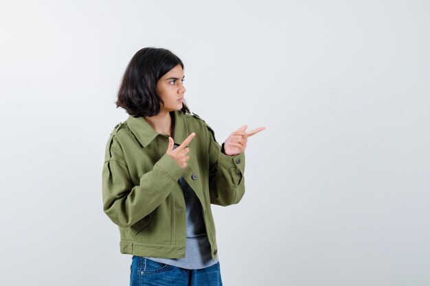 Niña apuntando hacia el lado derecho en abrigo, camiseta, jeans y luciendo confiada. vista frontal.