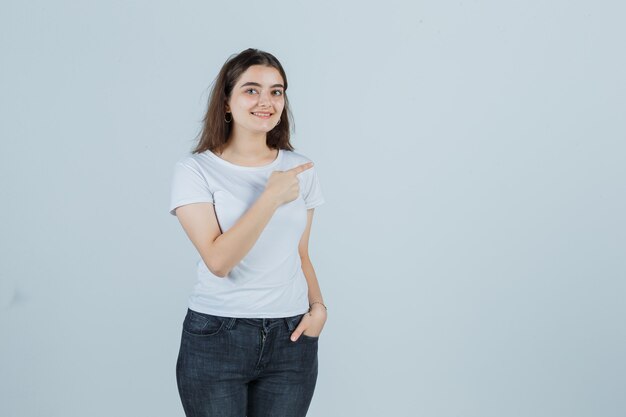 Niña apuntando hacia el lado derecho en camiseta, jeans y mirando feliz, vista frontal.