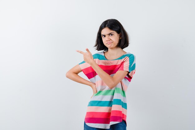 Niña apuntando hacia el lado izquierdo en camiseta y mirando dudosa. vista frontal.