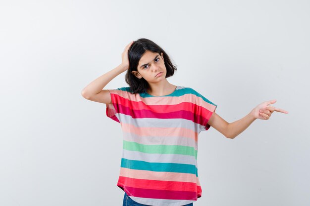 Niña apuntando a un lado, manteniendo la mano en la cabeza en la camiseta y mirando triste, vista frontal.