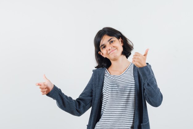 Niña apuntando a un lado, mostrando el pulgar hacia arriba en camiseta, chaqueta y luciendo alegre.