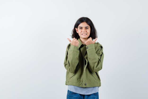 Niña apuntando a un lado con los pulgares en abrigo, camiseta, jeans y mirando feliz. vista frontal.