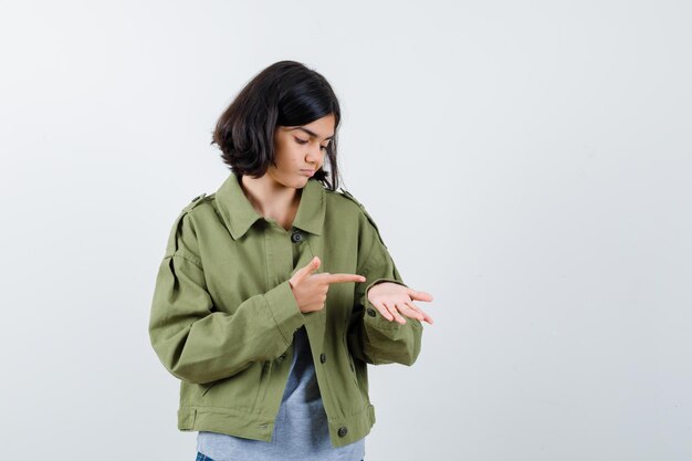 Niña apuntando a la mano con suéter gris, chaqueta caqui, pantalón de mezclilla y mirando enfocado, vista frontal.