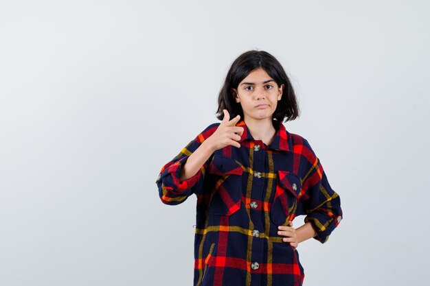 Niña apuntando mientras sostiene la mano en la cintura con camisa a cuadros y se ve linda, vista frontal.