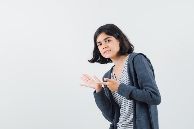 Niña apuntando a su palma vacía en camiseta, chaqueta y mirando pensativa.