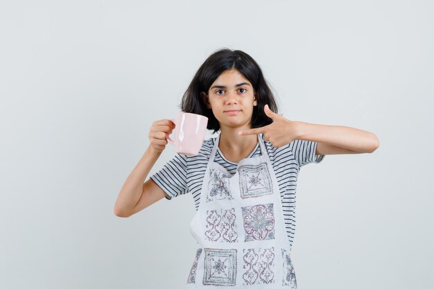 Niña apuntando a una taza de bebida en camiseta, delantal y mirando confiado,