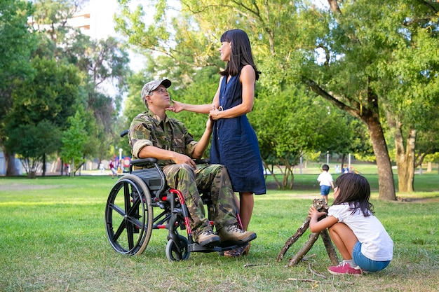 Niña arreglando leña para fogatas al aire libre mientras su mamá y papá militar discapacitado tomados de la mano y hablando. Veterano discapacitado o concepto de familia al aire libre