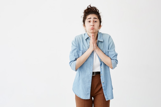 La niña arrepentida con el pelo oscuro en moño se ve sombría y desesperada, vestida con una camisa vaquera y pantalones marrones, mantiene las palmas juntas, suplica perdón, se siente culpable. Suplicando linda chica adorable