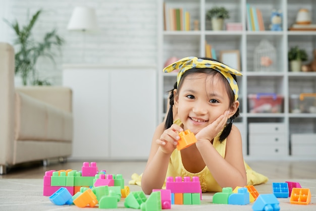 Niña asiática acostada en el piso en casa y jugando con coloridos bloques de construcción