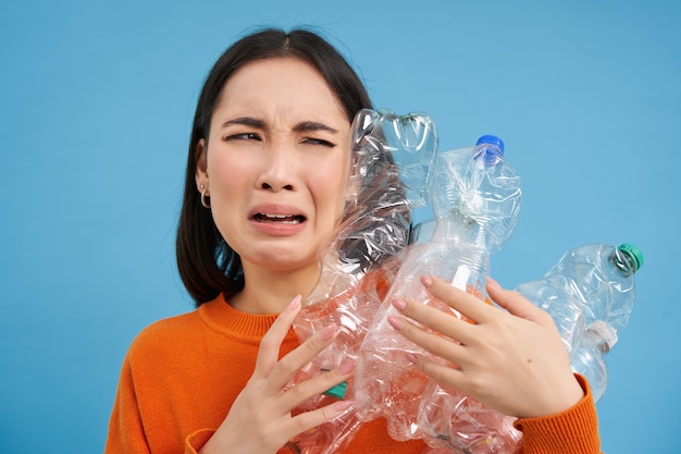 Niña asiática llorando sosteniendo botellas de plástico molesta reciclando de pie sobre fondo azul