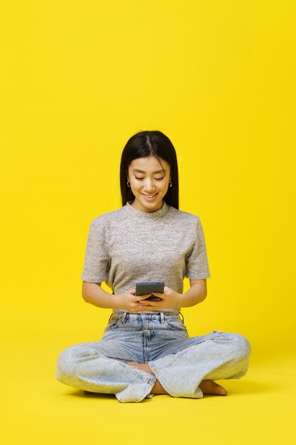 Niña asiática sentada en el suelo sosteniendo mensajes de texto de teléfonos inteligentes o comprando en línea aislada en fondo amarillo Colocación de productos para publicidad de aplicaciones móviles Copiar espacio