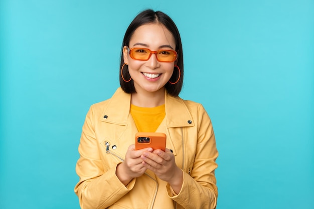 Niña asiática sonriente con gafas de sol usando una aplicación de teléfono inteligente sosteniendo un teléfono móvil de pie sobre fondo azul