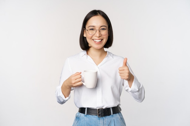 Niña asiática sonriente sosteniendo una taza blanca y pulgares arriba recomendando beber café o té de pie sobre fondo blanco