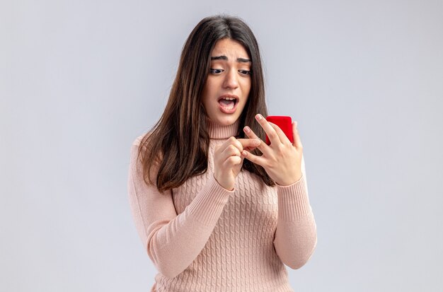 Niña asustada en el día de San Valentín sosteniendo y mirando el anillo de bodas aislado sobre fondo blanco.