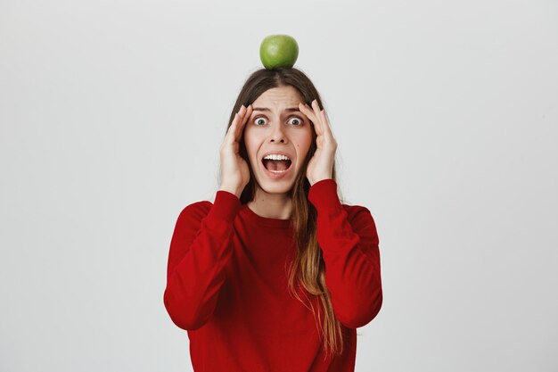 Niña asustada grita de pánico al ver una flecha voladora, sosteniendo el objetivo de manzana en la cabeza