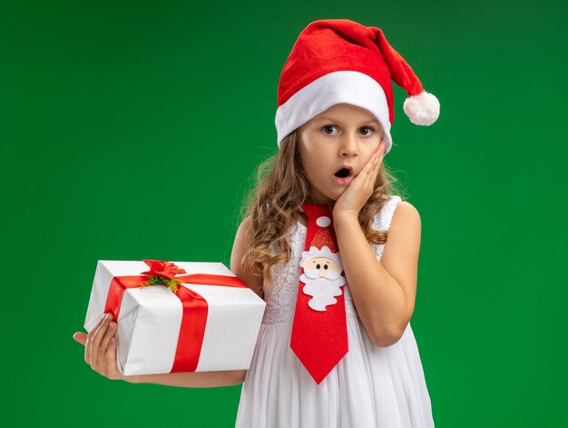 Niña asustada vistiendo gorro de navidad con corbata sosteniendo caja de regalo poniendo la mano en la mejilla aislado en la pared verde