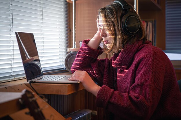 Una niña con auriculares detrás de una computadora portátil en la luz del sol a través de las persianas.