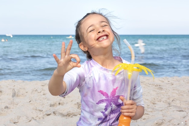 La niña bebe jugo sentada en la arena cerca del mar