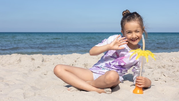 La niña bebe jugo sentada en la arena cerca del mar