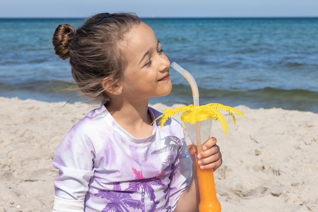 La niña bebe jugo sentada en la arena cerca del mar